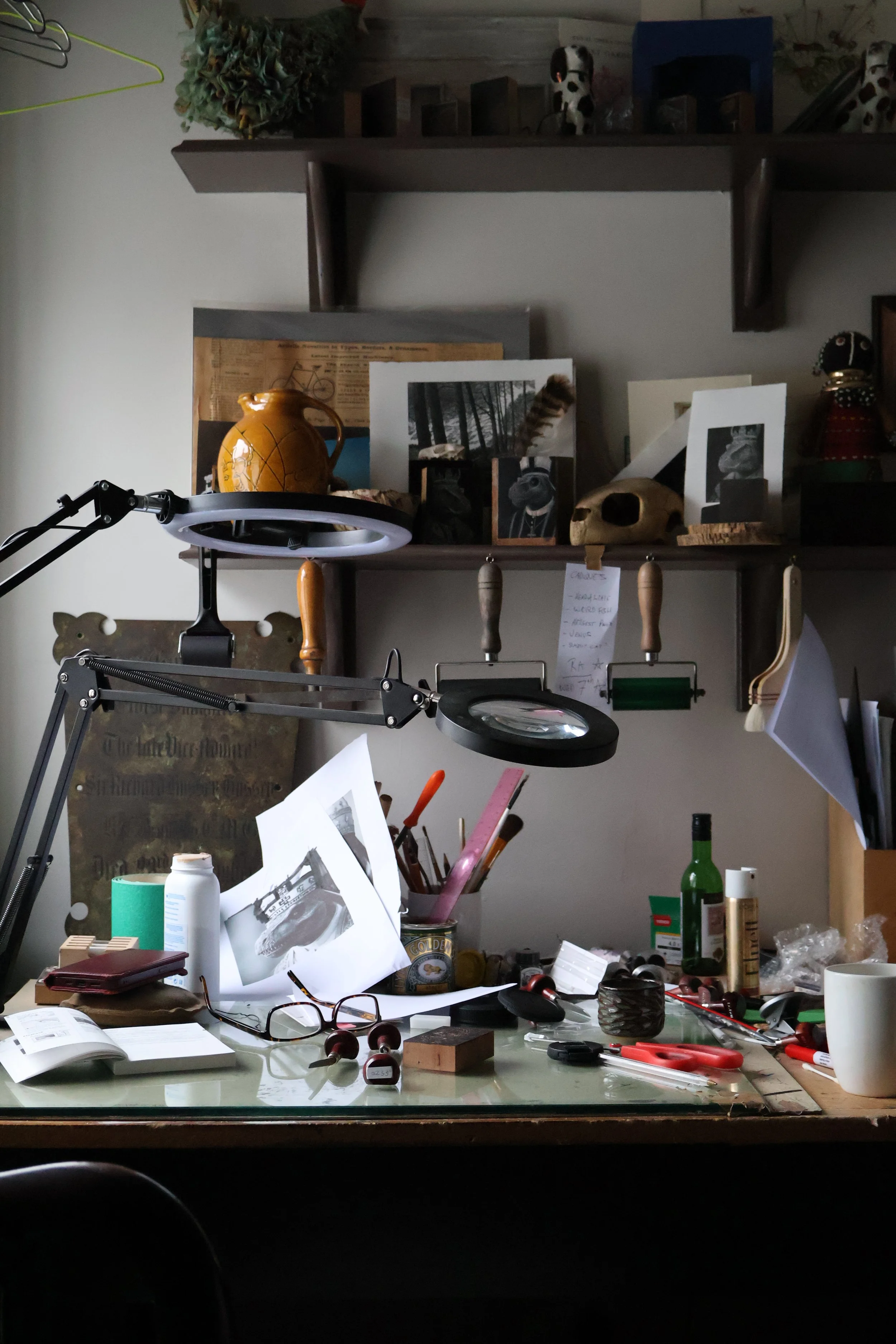 A cluttered desk with papers, glasses, and art supplies, with shelves above holding photographs, a skull, and figurines.