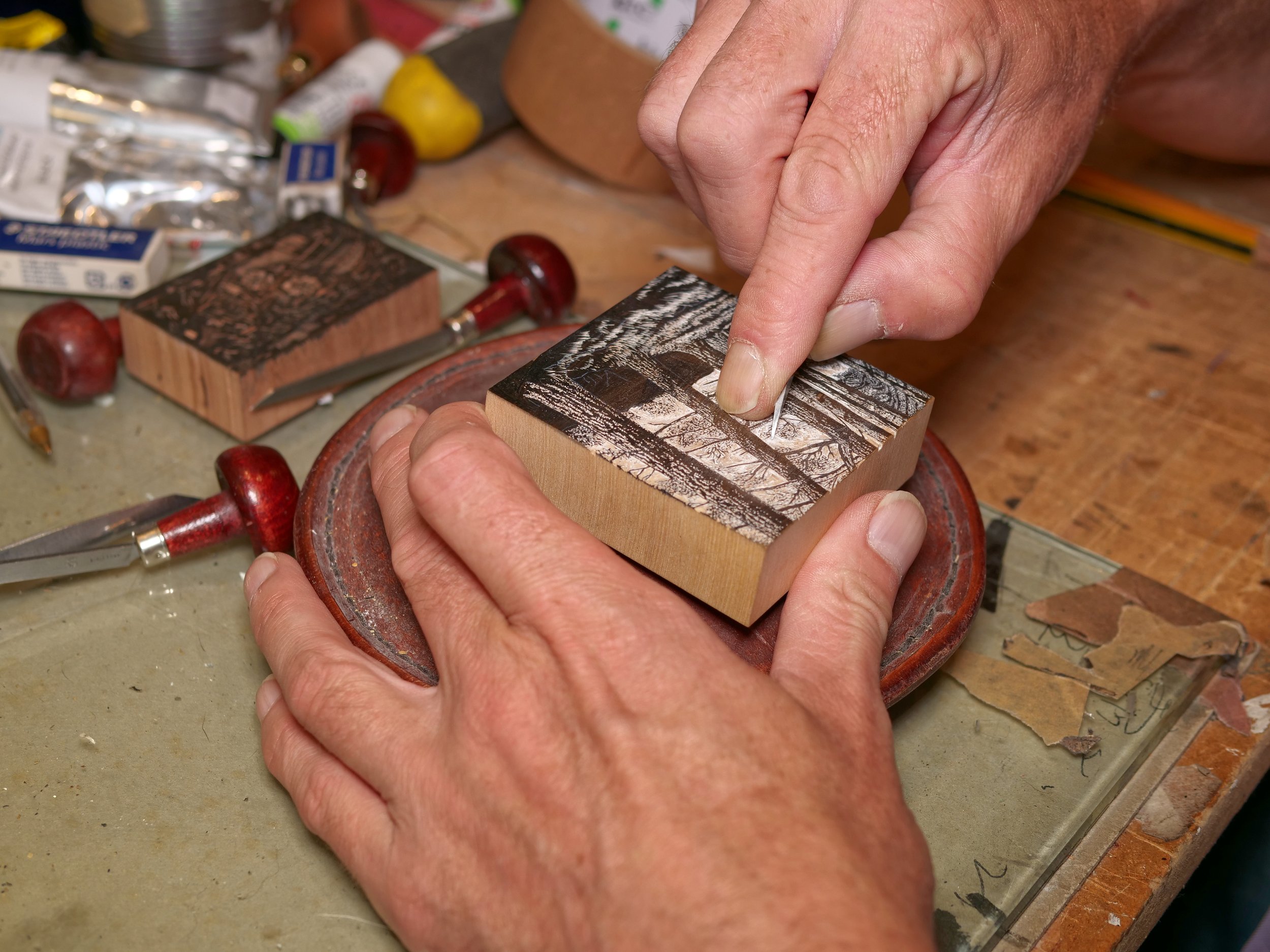 A person is carving a design into a wooden block with a small metal tool. The workspace has various woodworking stamps, tools, and supplies scattered around.