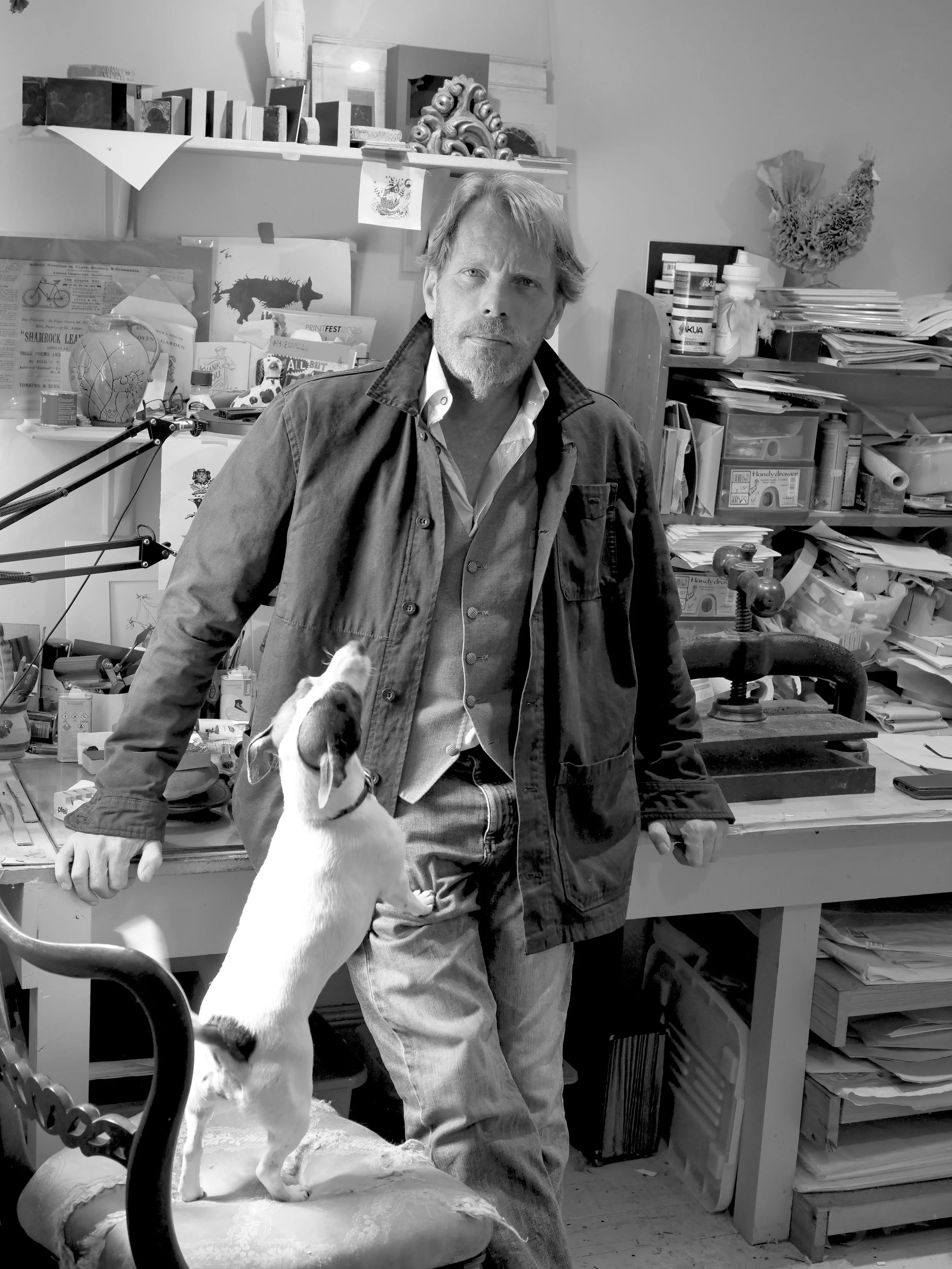 A man in a dark jacket standing in a cluttered workspace with a puppy standing on a chair, looking up at him.
