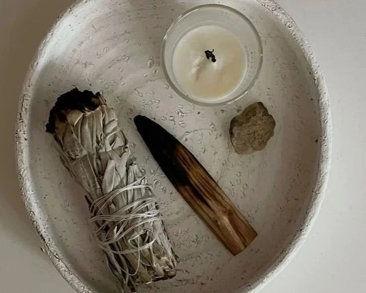 A white bowl holding a bundle of sage smudging sticks, a wooden stick, a small candle, and a small rock.