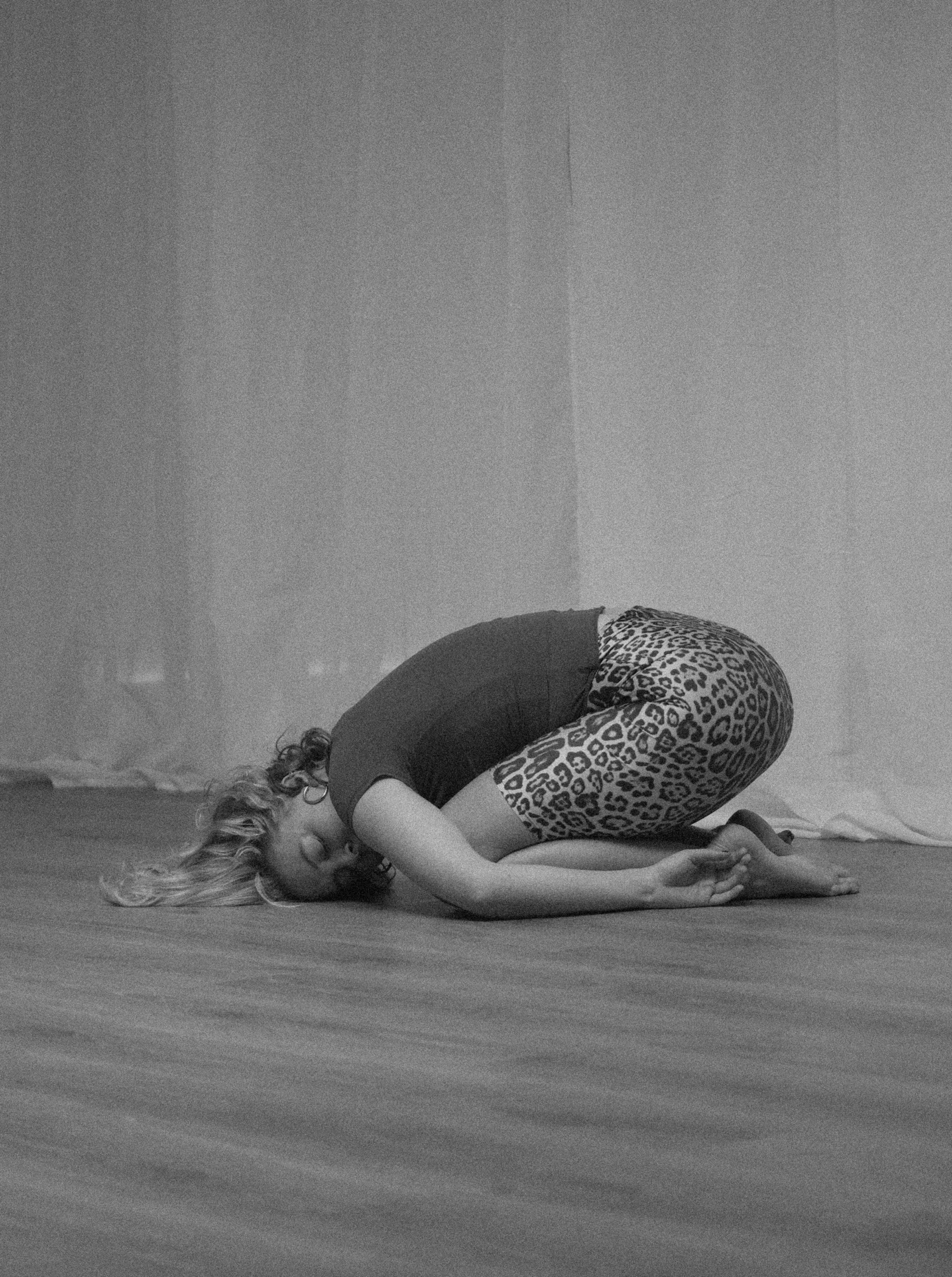 A woman with curly hair practicing yoga in a child's pose on a wooden floor, wearing leopard print leggings and a black top, in front of a plain wall.