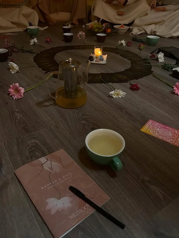 A table set for a gathering with a wooden centerpiece decorated with flowers, cups of tea or coffee, a pitcher of tea, and a lit candle. There are people sitting around the table, some with their hands visible. A brochure and a card are also on the table.