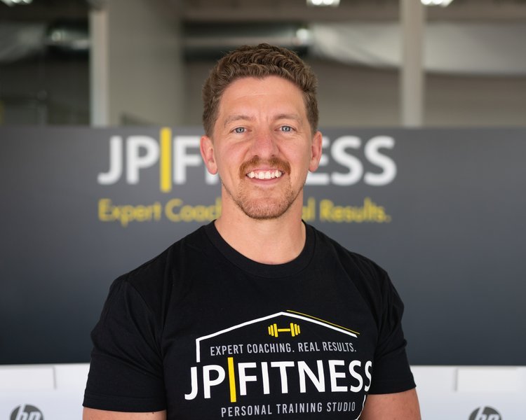 A man with short curly hair and a beard, smiling, wearing a black T-shirt with the words 'JP Fitness' and a barbell graphic, standing in front of a dark background with the same logo.