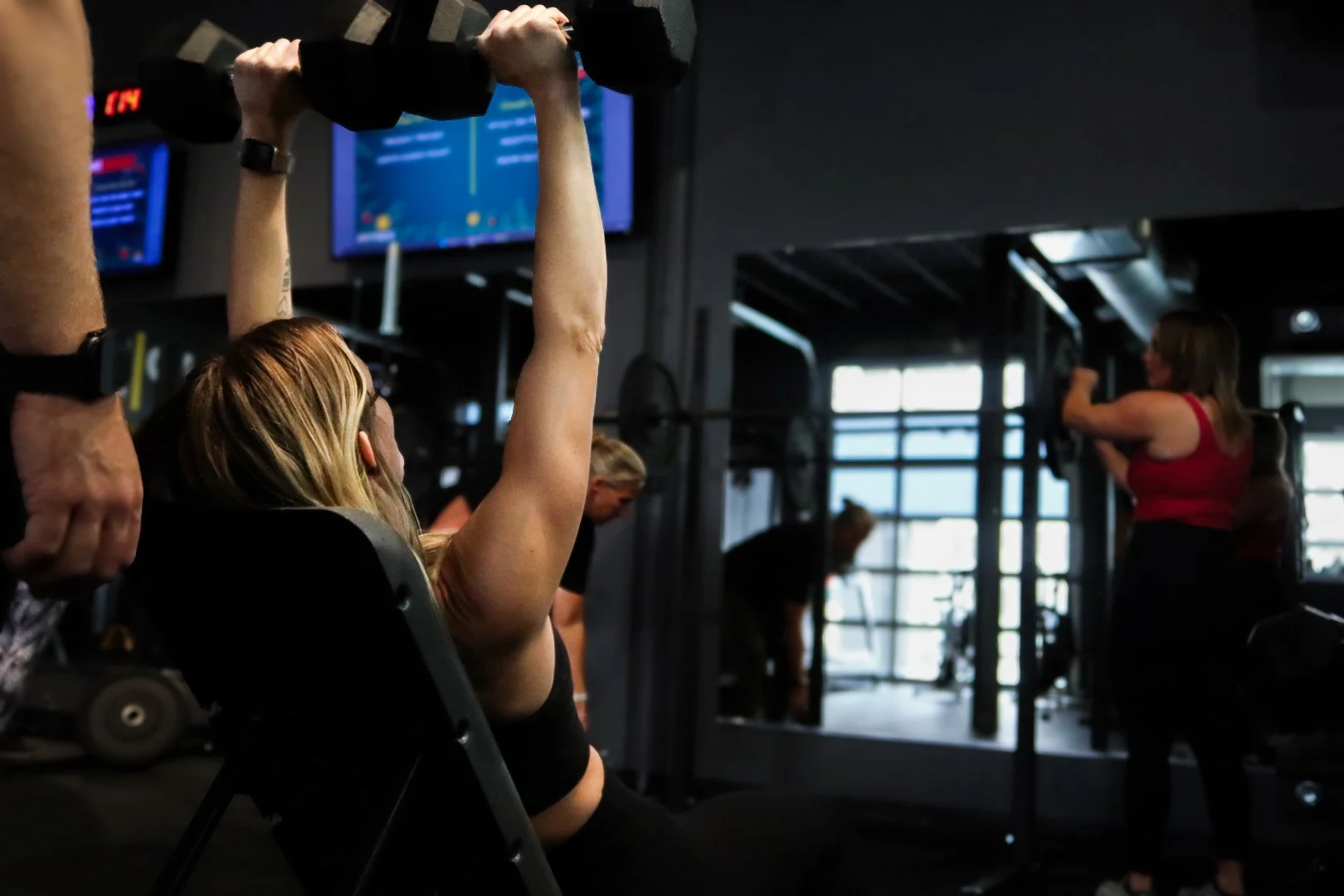 A woman is exercising with dumbbells in a gym, lifting them above her head while sitting in a chair. Other people are working out in the background, including a woman near a mirror and a person bent over. The gym has dark lighting with windows letting in natural light, and screens displaying information on the wall.