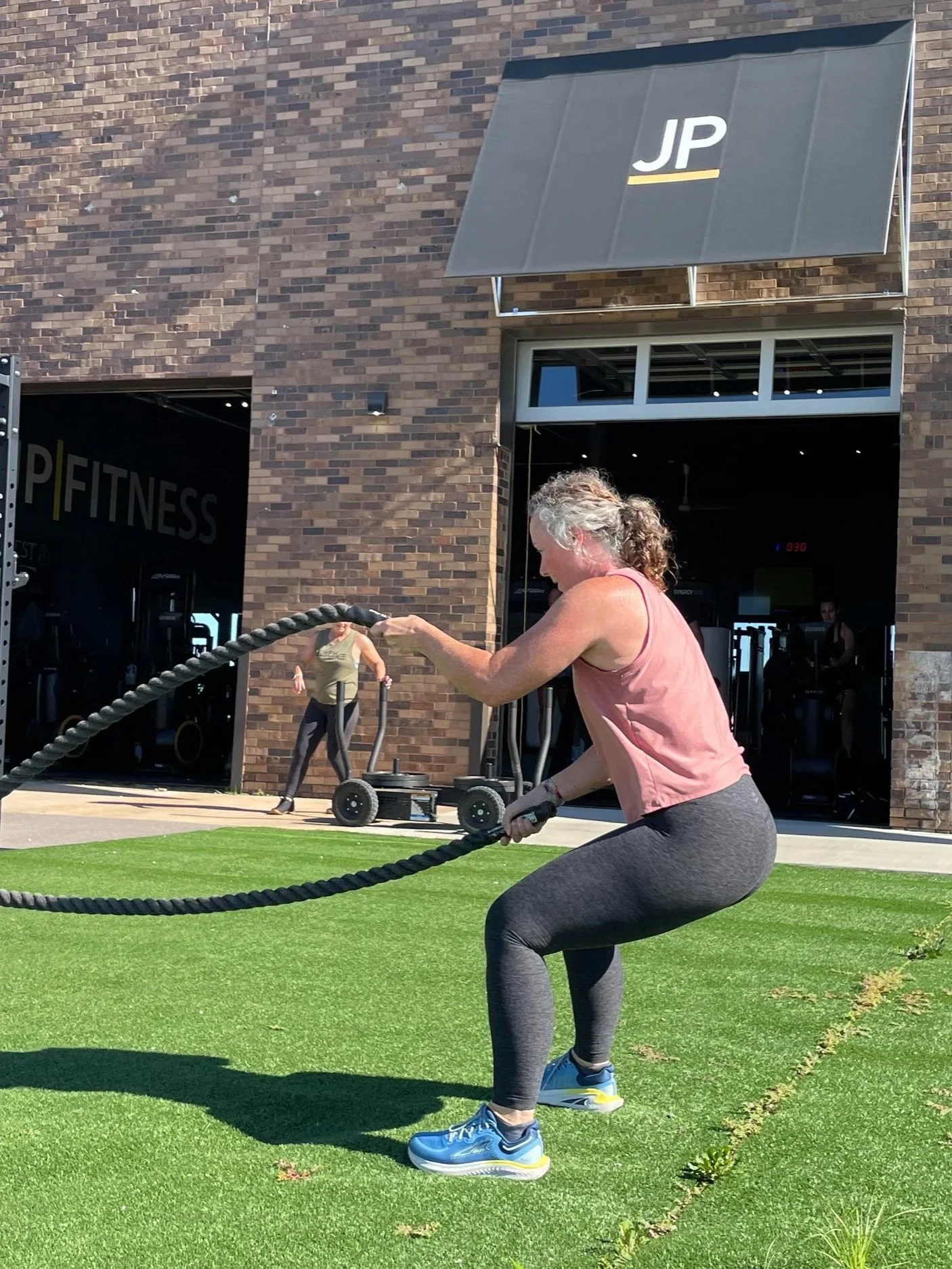 Woman exercising with battle ropes outside a gym with a brick exterior and open doors.