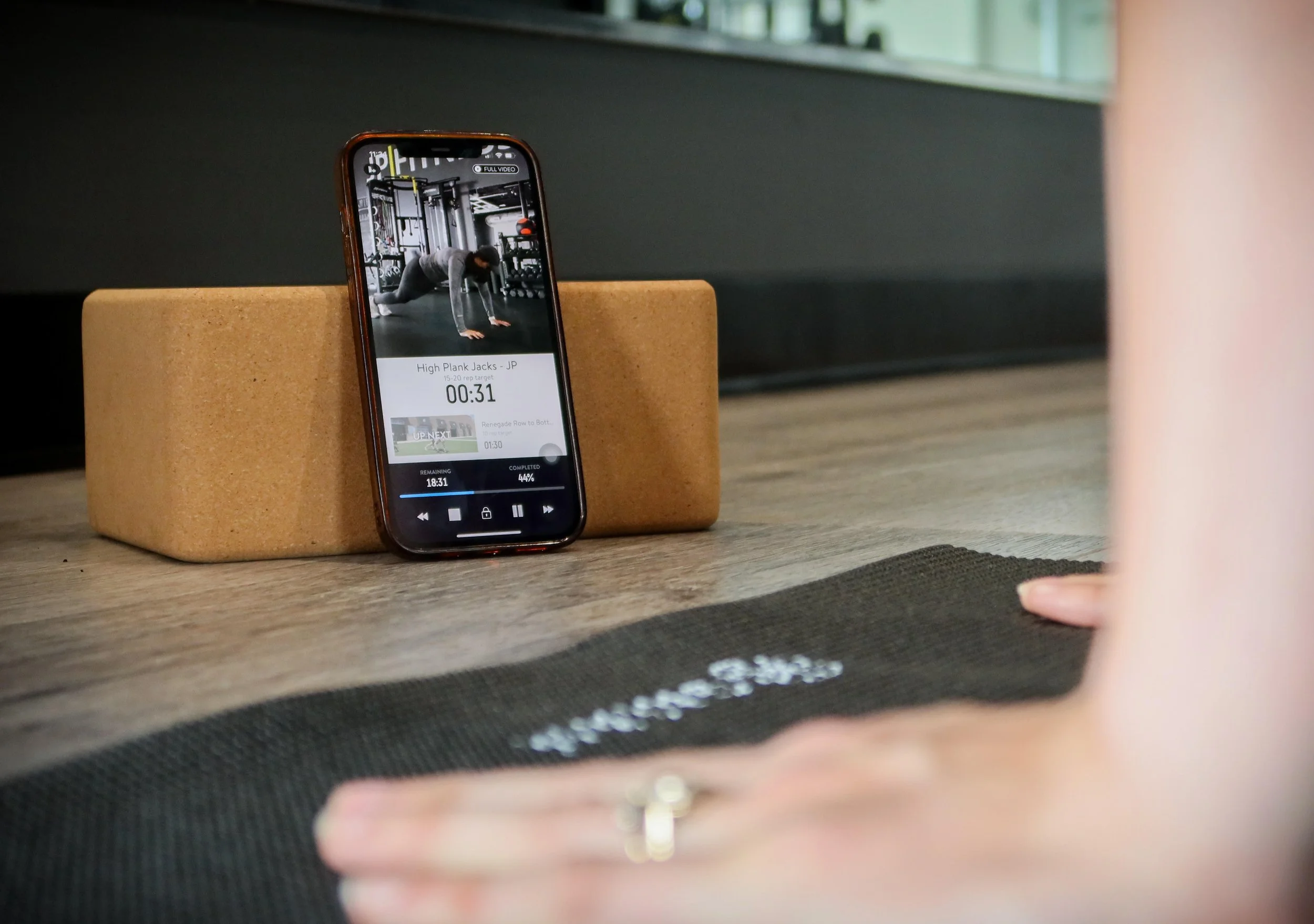 Person practicing yoga in a gym, with a smartphone showing a workout video placed on a wooden block on the floor.