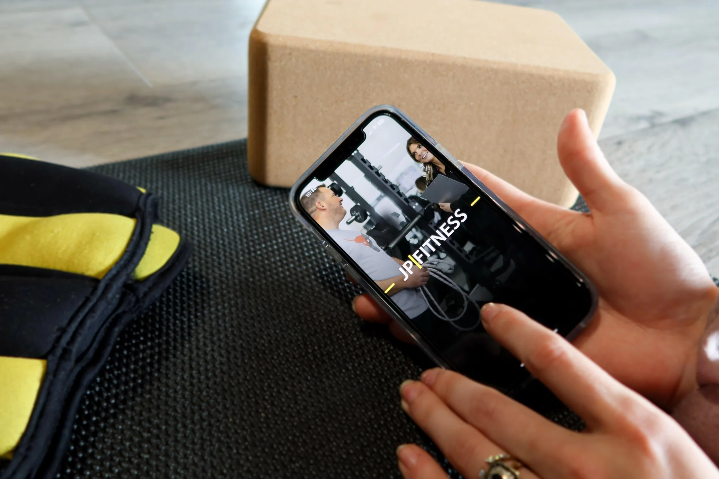 Person holding a smartphone with JD Fitness app open, sitting at a table with a black textured surface, yellow and black fitness equipment, and a brown foam block in the background.