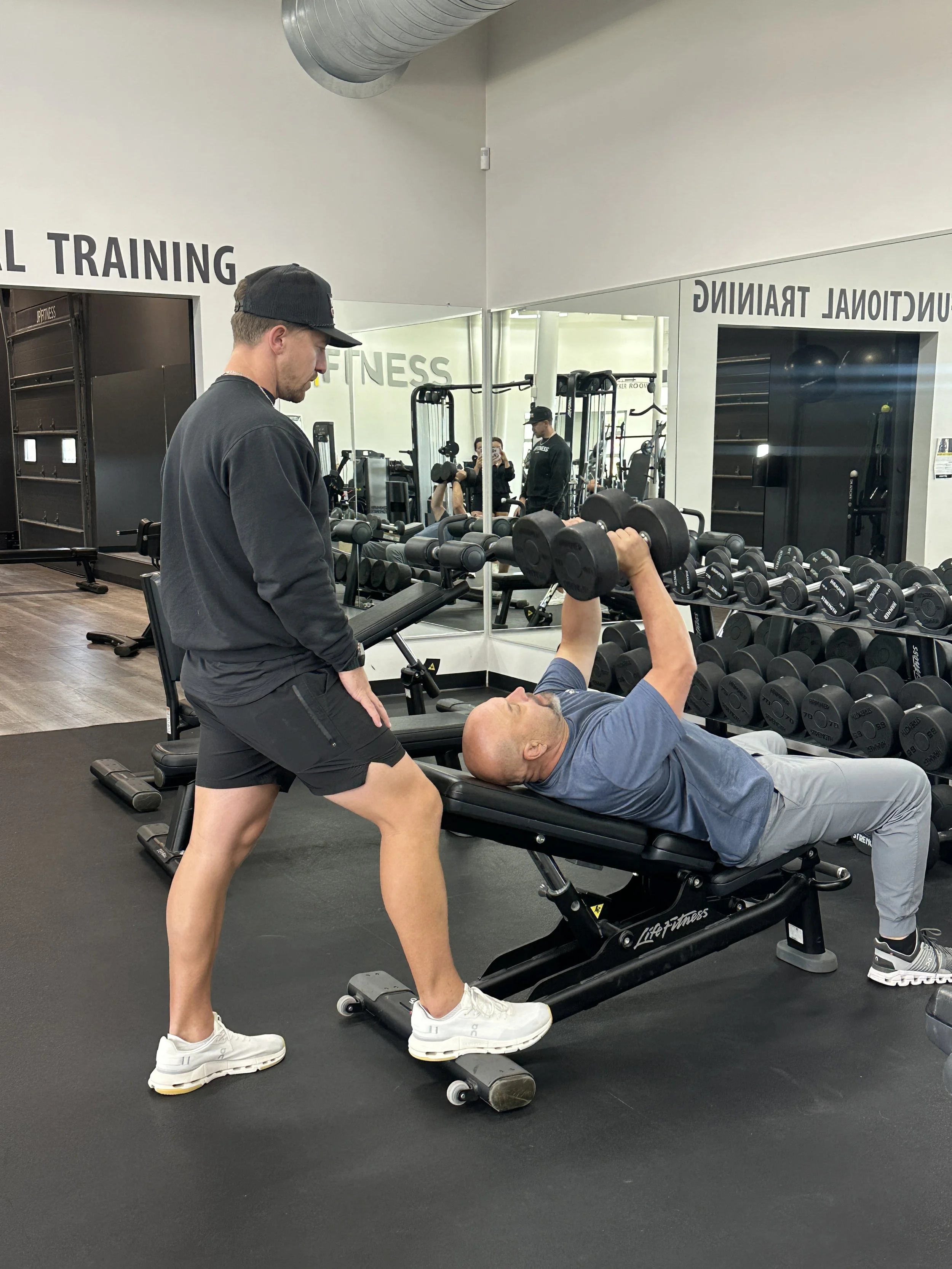 A personal trainer gives advice to a man lifting a barbell in a gym with a sign that reads 'FUNCTIONAL TRAINING' in the background.