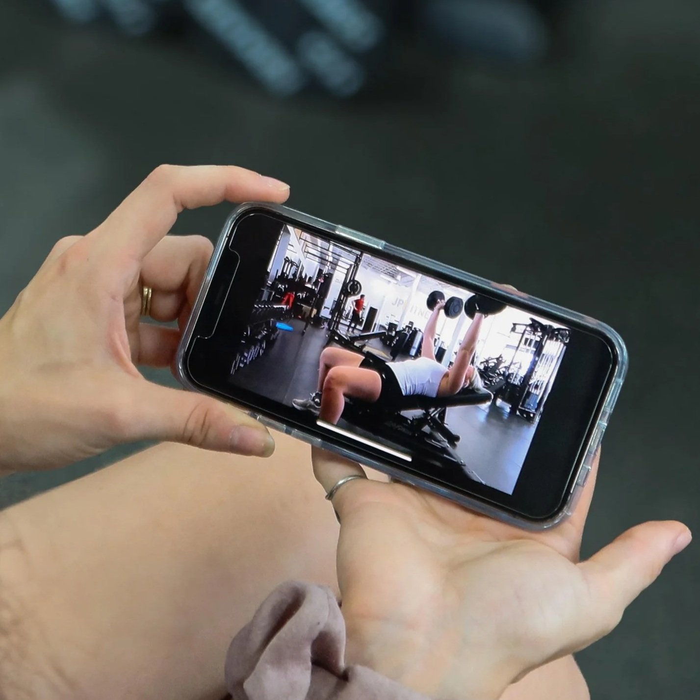 Person holding smartphone taking a photo of a woman doing a bench press at a gym.