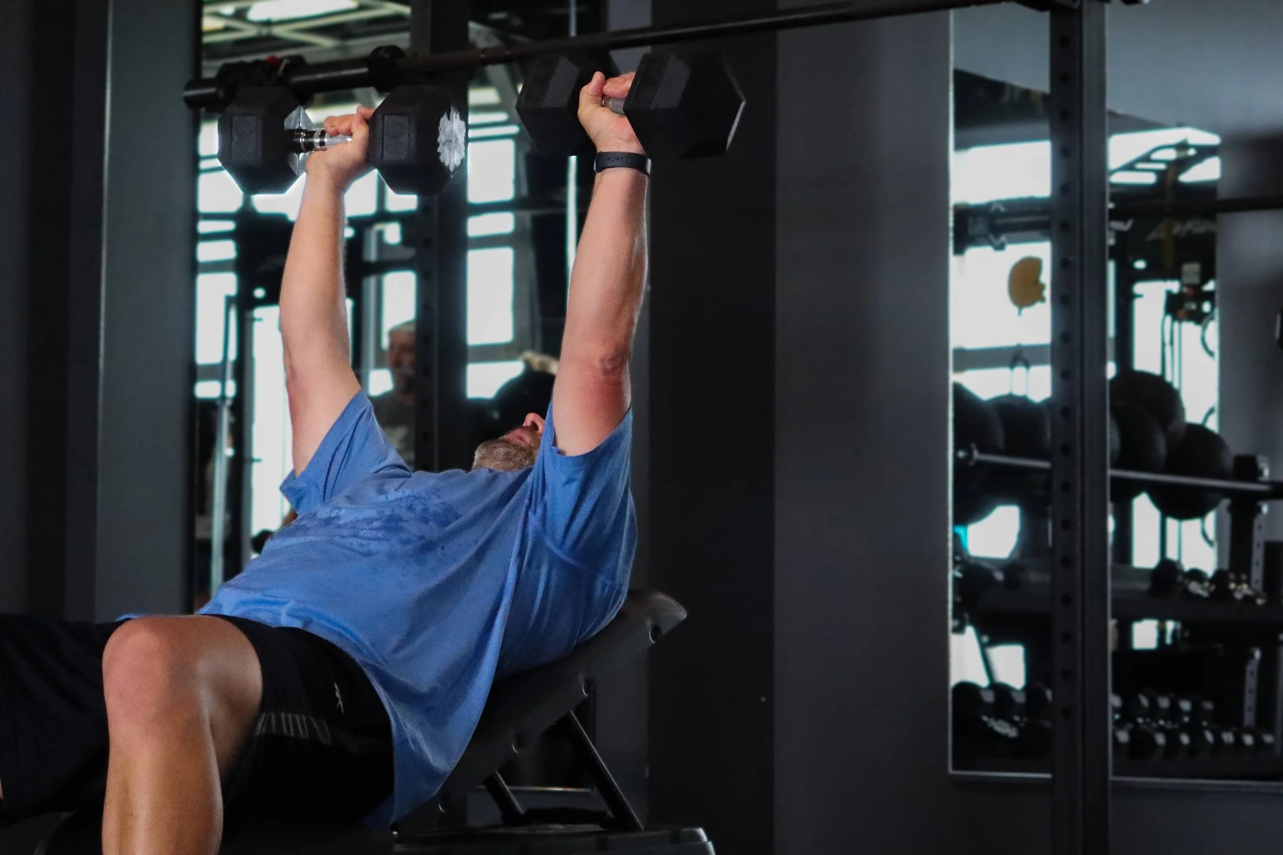 A man in a blue athletic shirt and black shorts is lying on a workout bench, lifting a black dumbbell with both hands in a gym.
