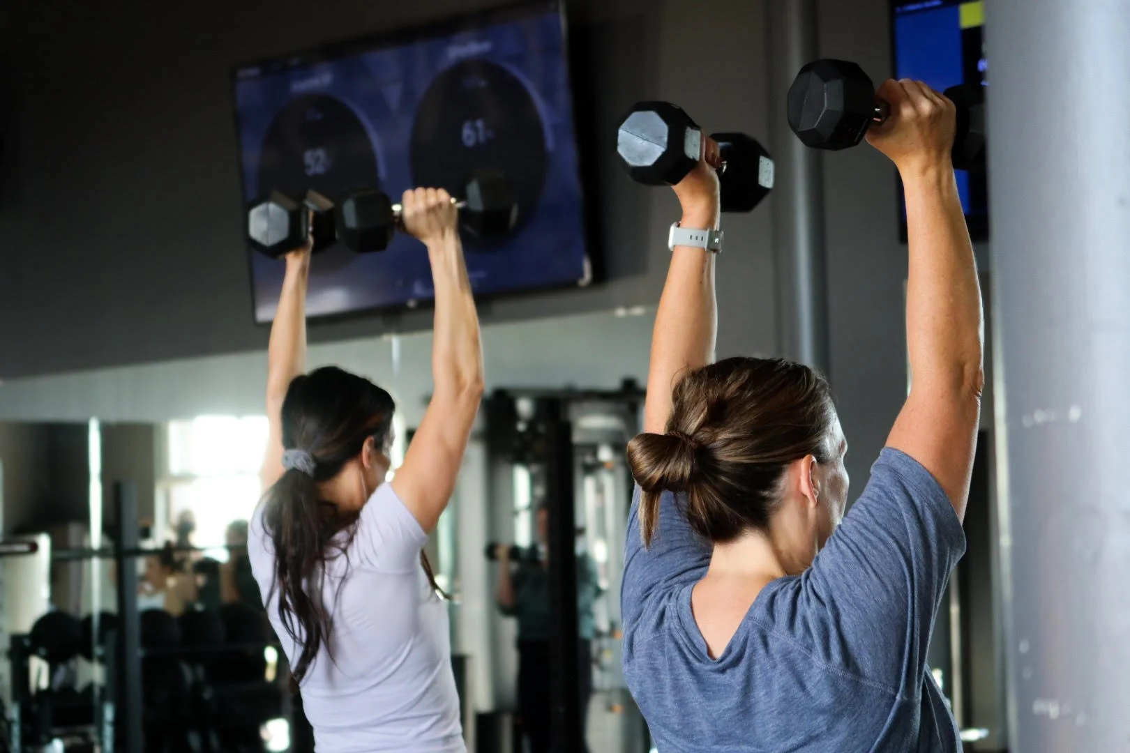 Two women lifting dumbbells during a workout class in a gym with monitors in the background.