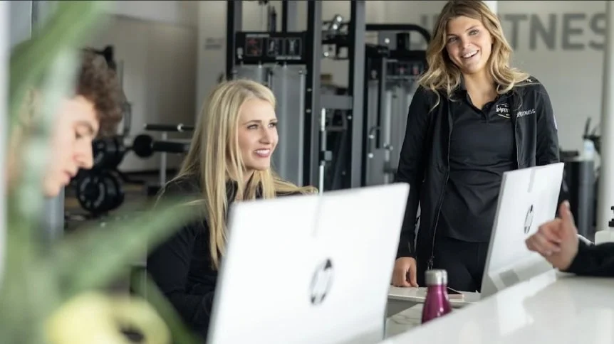 Three women in a gym office, smiling and talking, with fitness equipment in the background and computers on the table.
