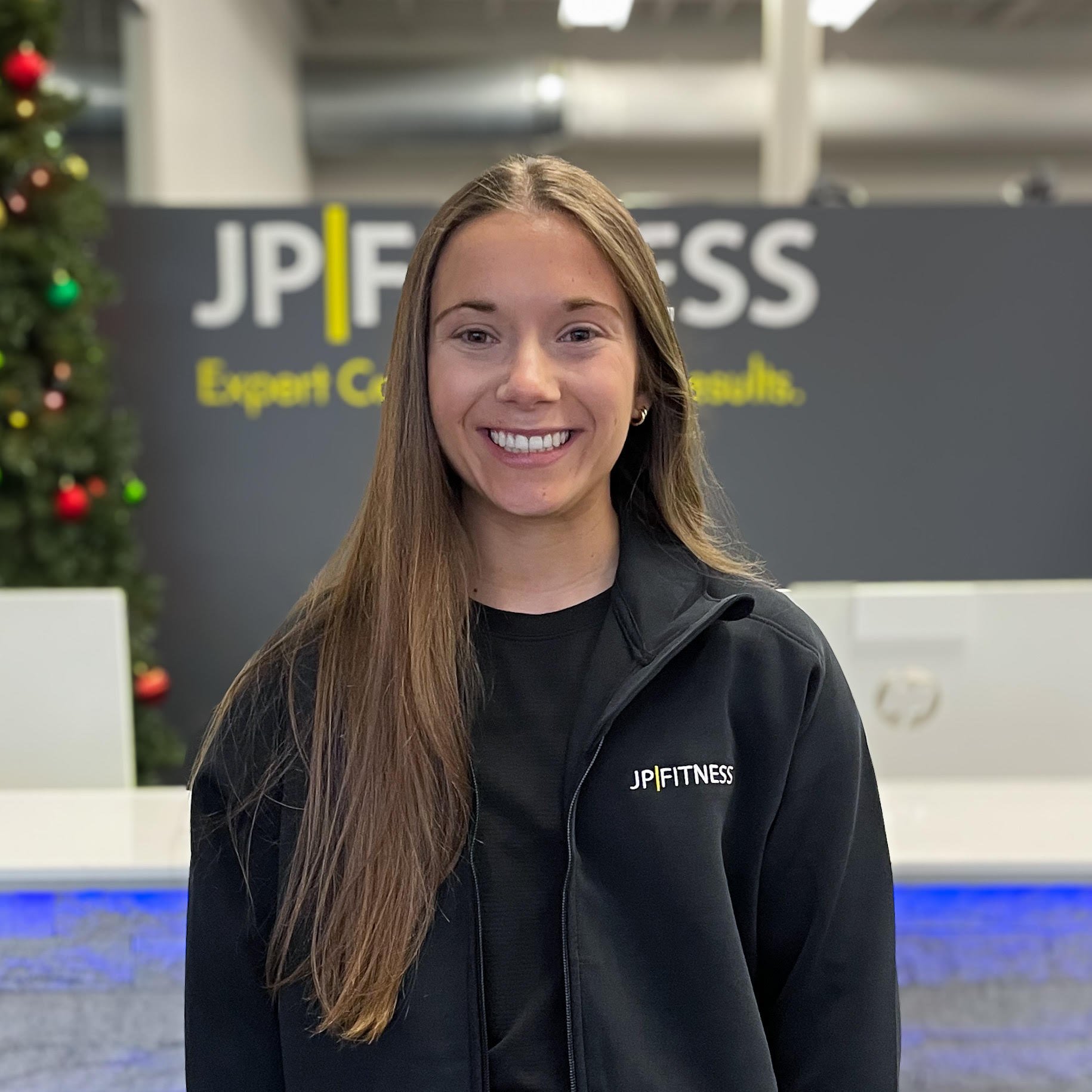 A woman with long brown hair smiling while standing in front of a JP Fitness reception area with a Christmas tree to the side.