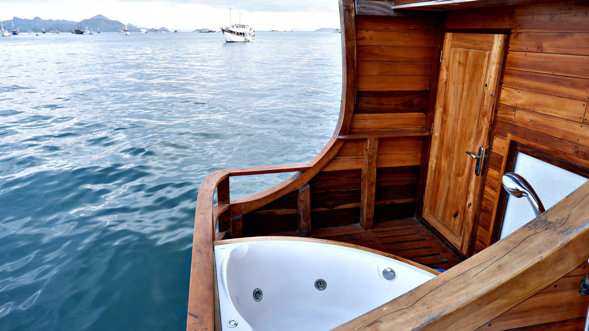 A wooden boat with a built-in jacuzzi overlooking the water with sailboats and mountains in the distance.