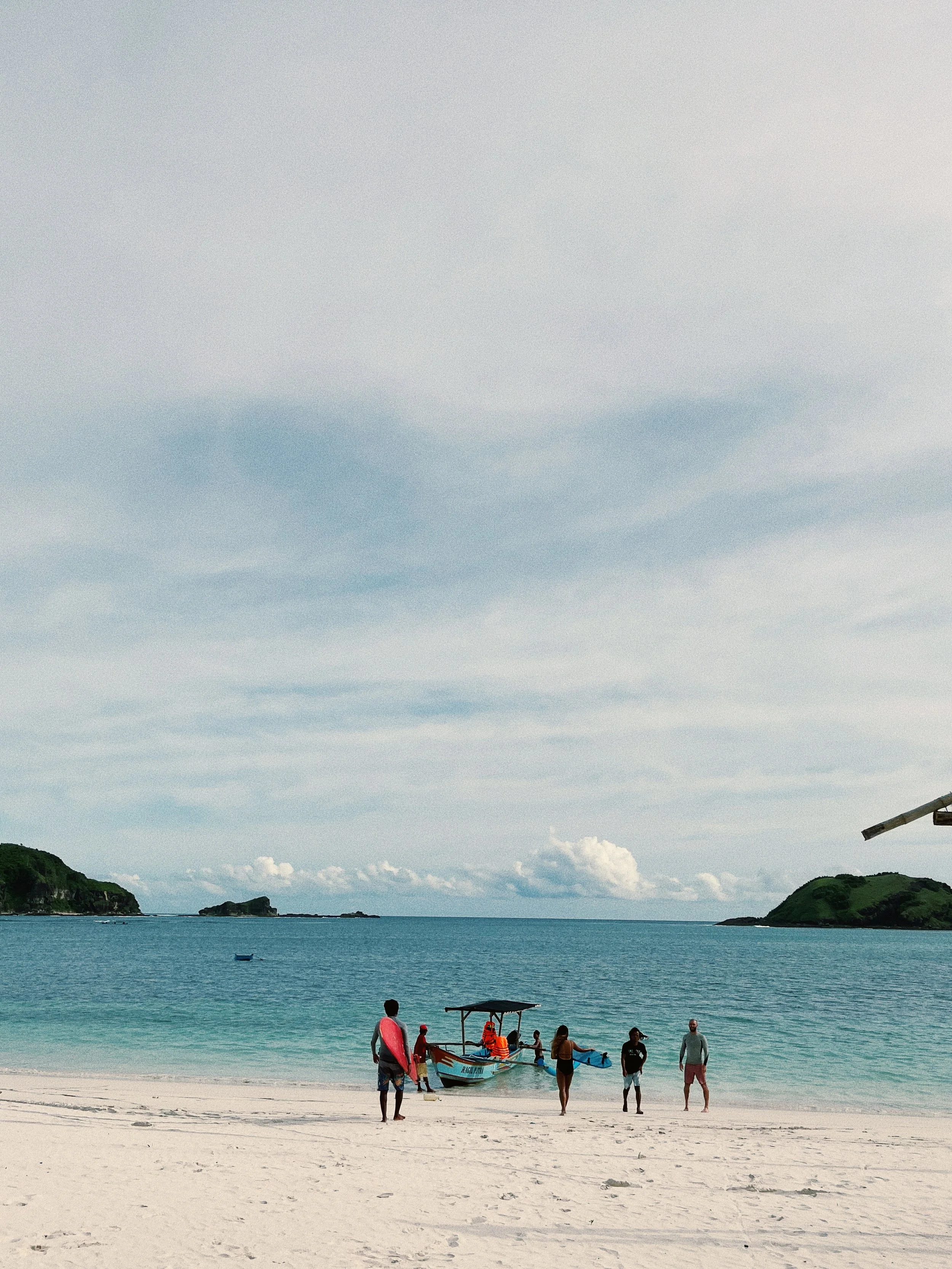 People on a beach near a small boat docked on the shore, with a background of clear blue water and small islands.