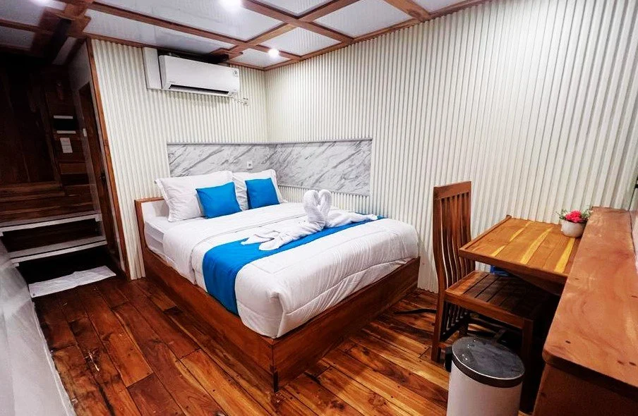 A cozy hotel room featuring a bed with white linens and blue accents, a towel folded into a swan shape on the bed, a small wooden desk with a flower pot, a wooden chair, wooden flooring, a marble-patterned headboard, and an air conditioning unit on t