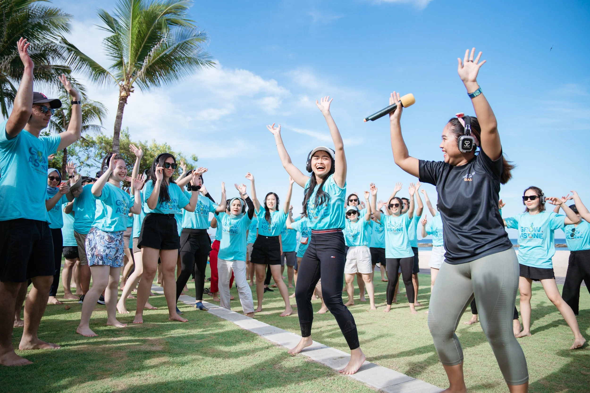 Group of people outdoors, participating in a dance or exercise activity on a sunny day, with palm trees and a blue sky in the background.