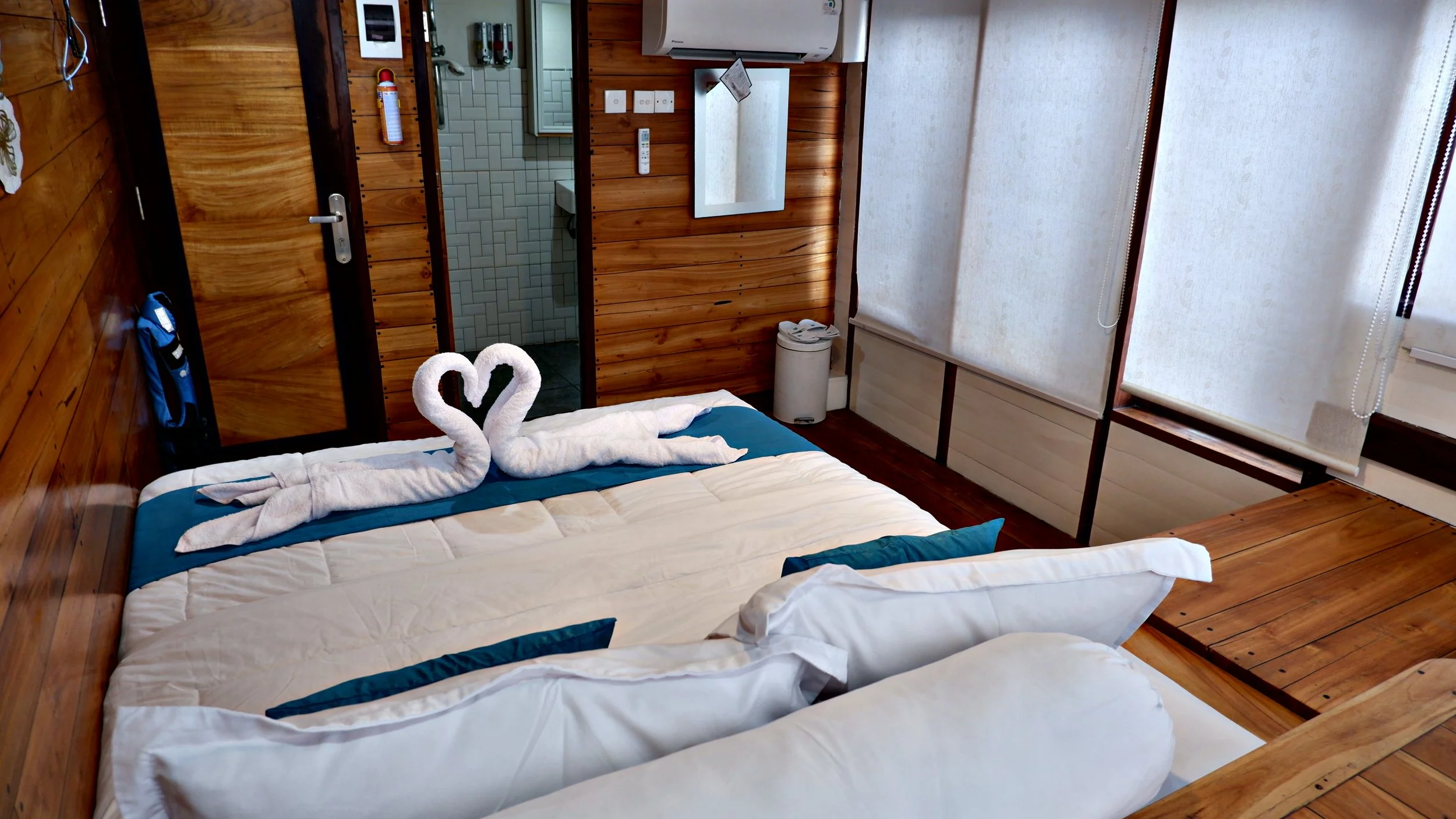 A cozy hotel room with a bed featuring folded towels shaped like swans forming a heart, a wooden desk, and a view of the bathroom with a shower visible through an open door. The room has wood-paneled walls and rolled-up blinds covering the windows.