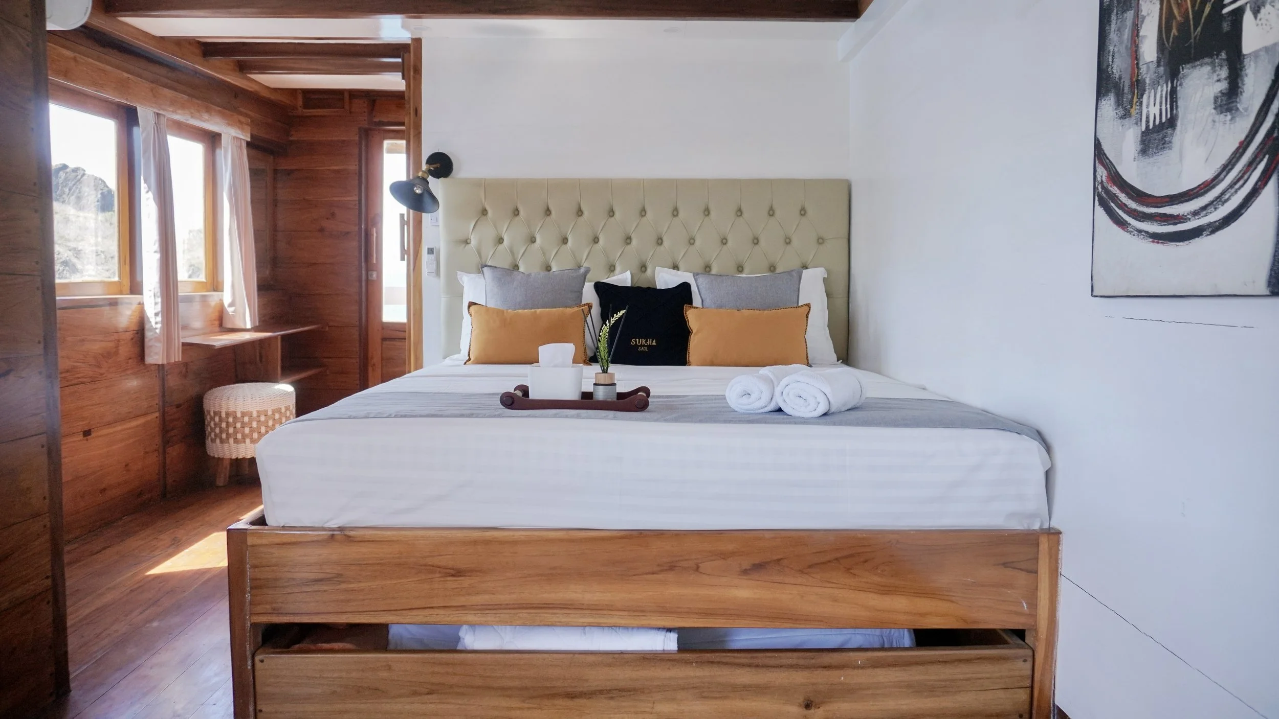 A cozy bedroom with wooden walls and floor, featuring a large bed with a beige tufted headboard, gray and mustard pillows, a tray with tissues and a small plant, with white towels on the bed. There's a window and door on the left, a bedside light, an