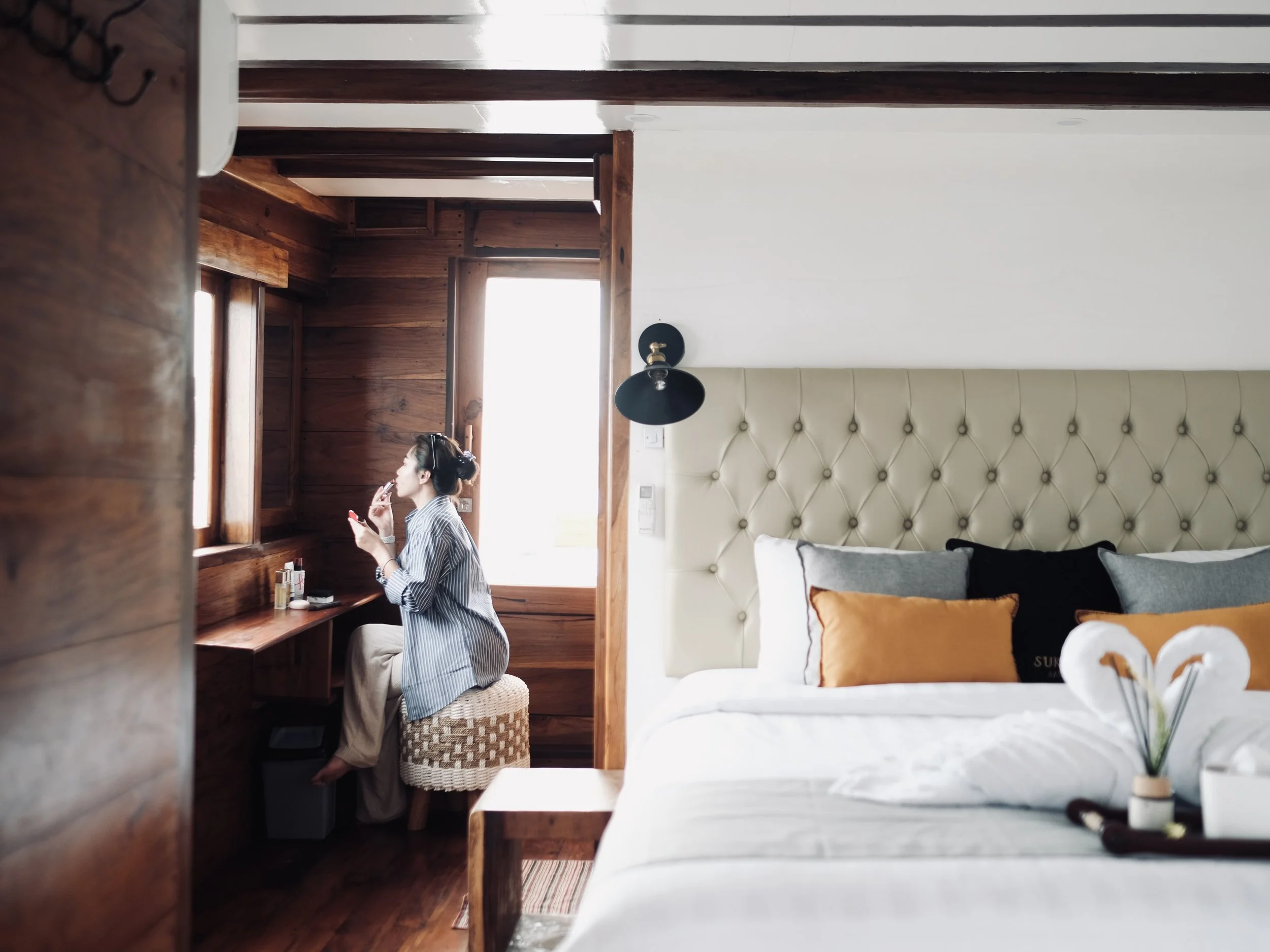 Woman sitting on a stool applying makeup in a hotel room with wood-paneled walls and a large bed with decorative pillows.