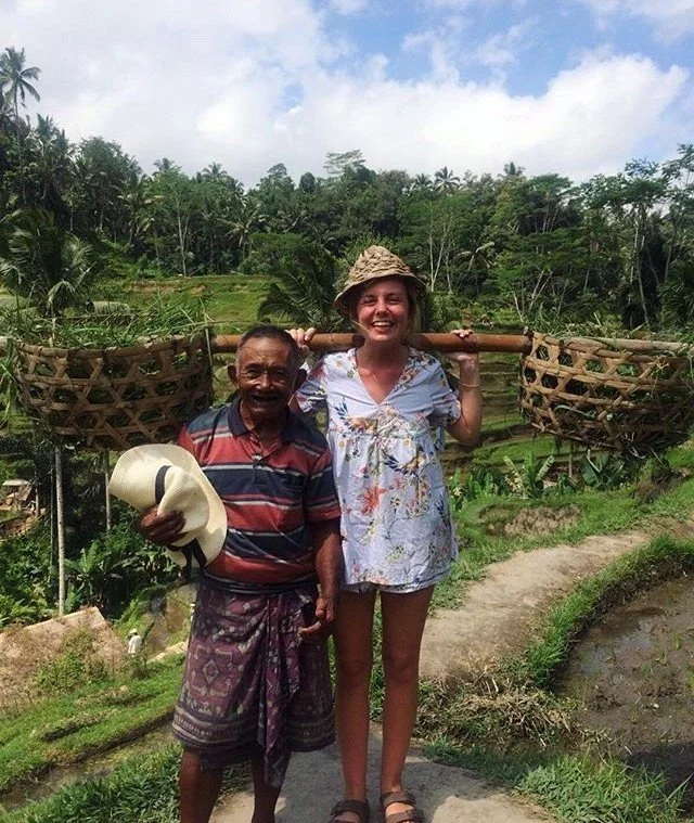 Two people in a lush, rural landscape. One person is wearing a patterned shirt and holding a hat, while the other is wearing a floral outfit and a straw hat, holding a large carrier across their shoulders. The background features green fields and palm trees.