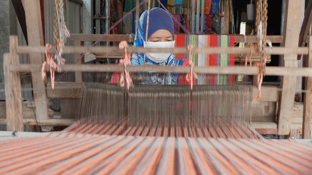 Person weaving fabric on a loom, wearing a blue headscarf and face mask, with colorful textiles in the background.