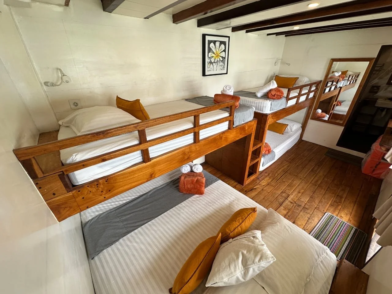 Room with wooden bunk beds, white bedding, and orange pillows, with a small bathroom mirror and a framed flower picture on the wall.