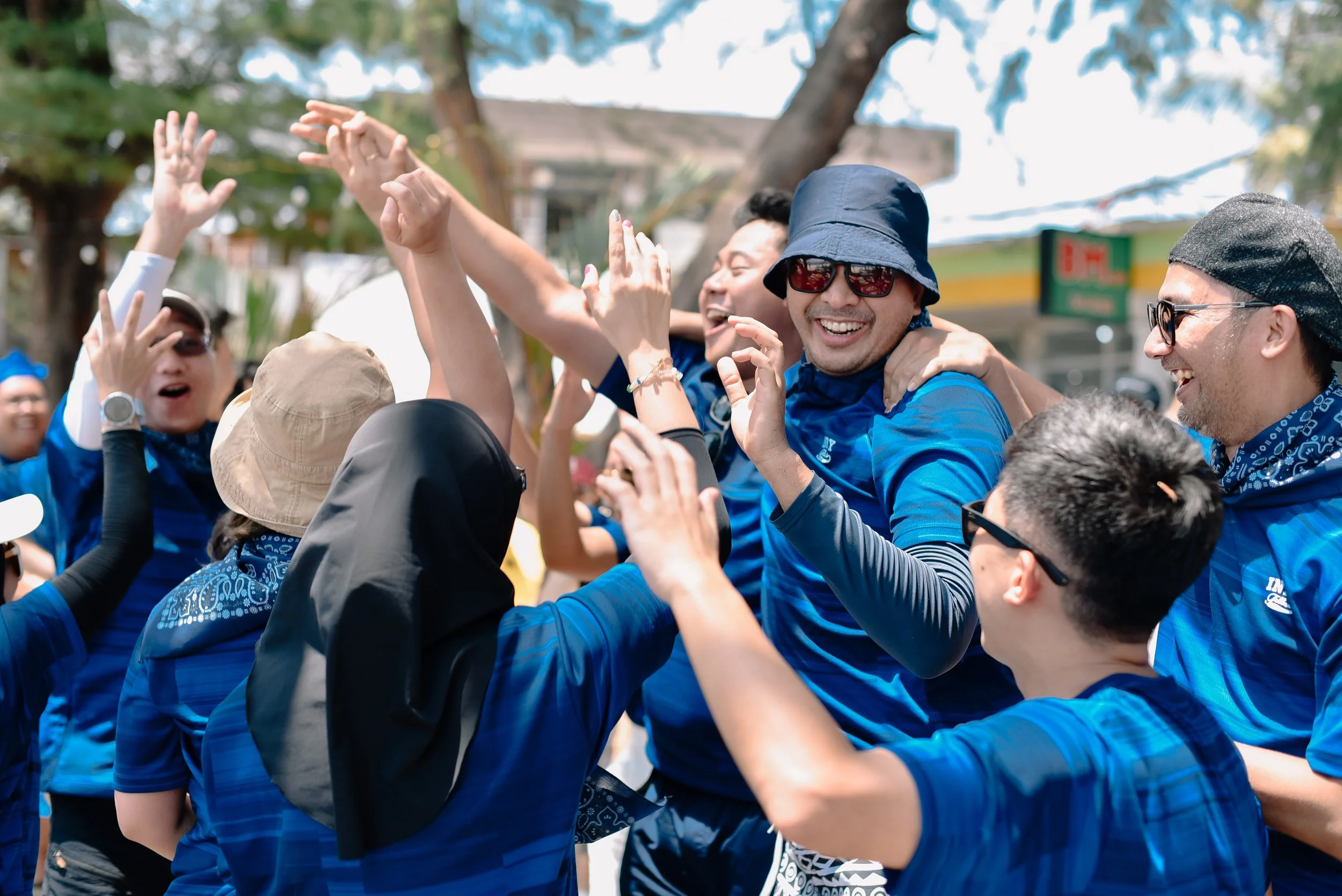 Group of people wearing blue shirts celebrating outdoors, smiling, and raising hands, with trees and a building in the background.
