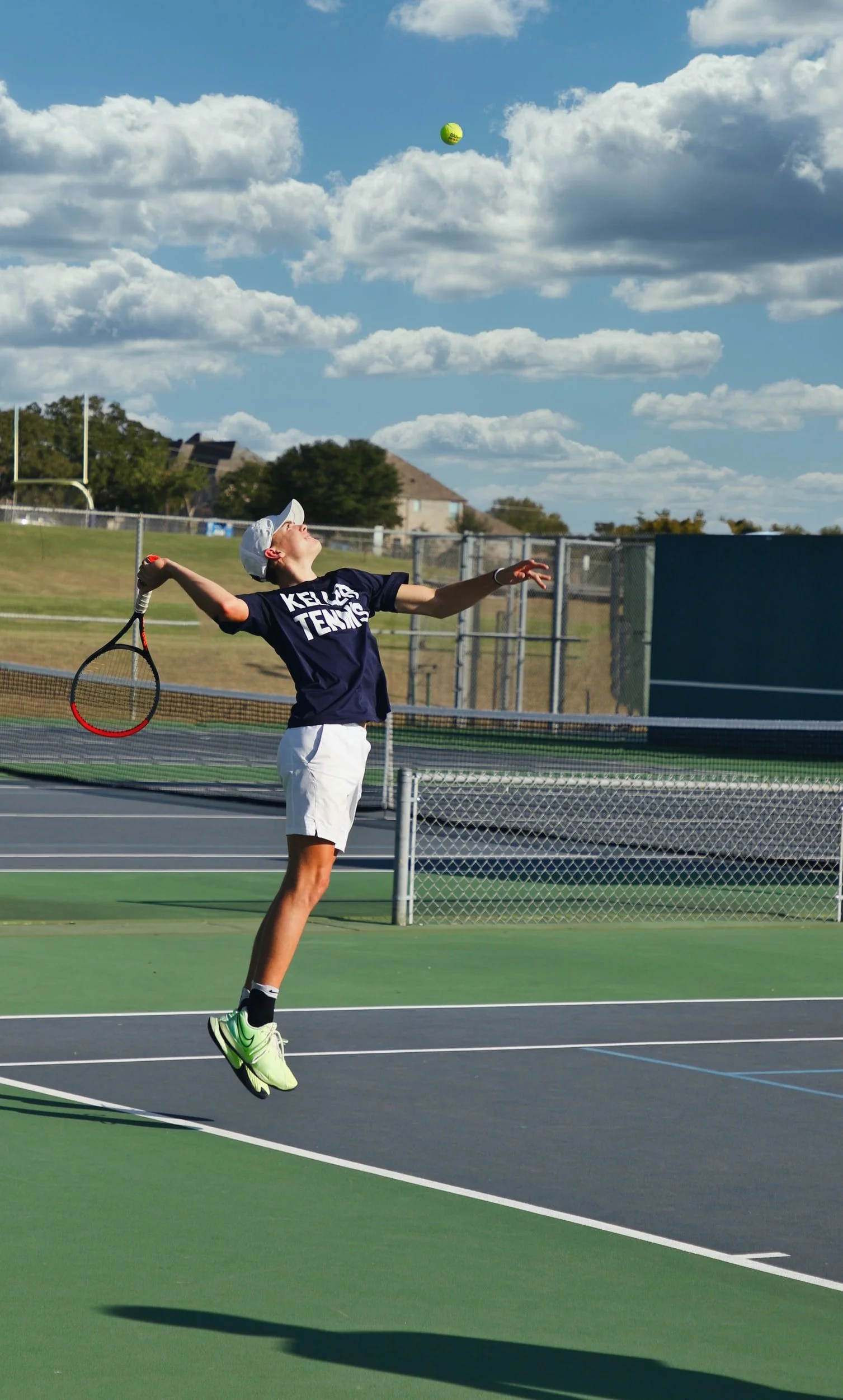 A young male tennis player in a navy T-shirt and white shorts leaps into the air on a tennis court to serve with a tennis racket, under a sky with scattered clouds.