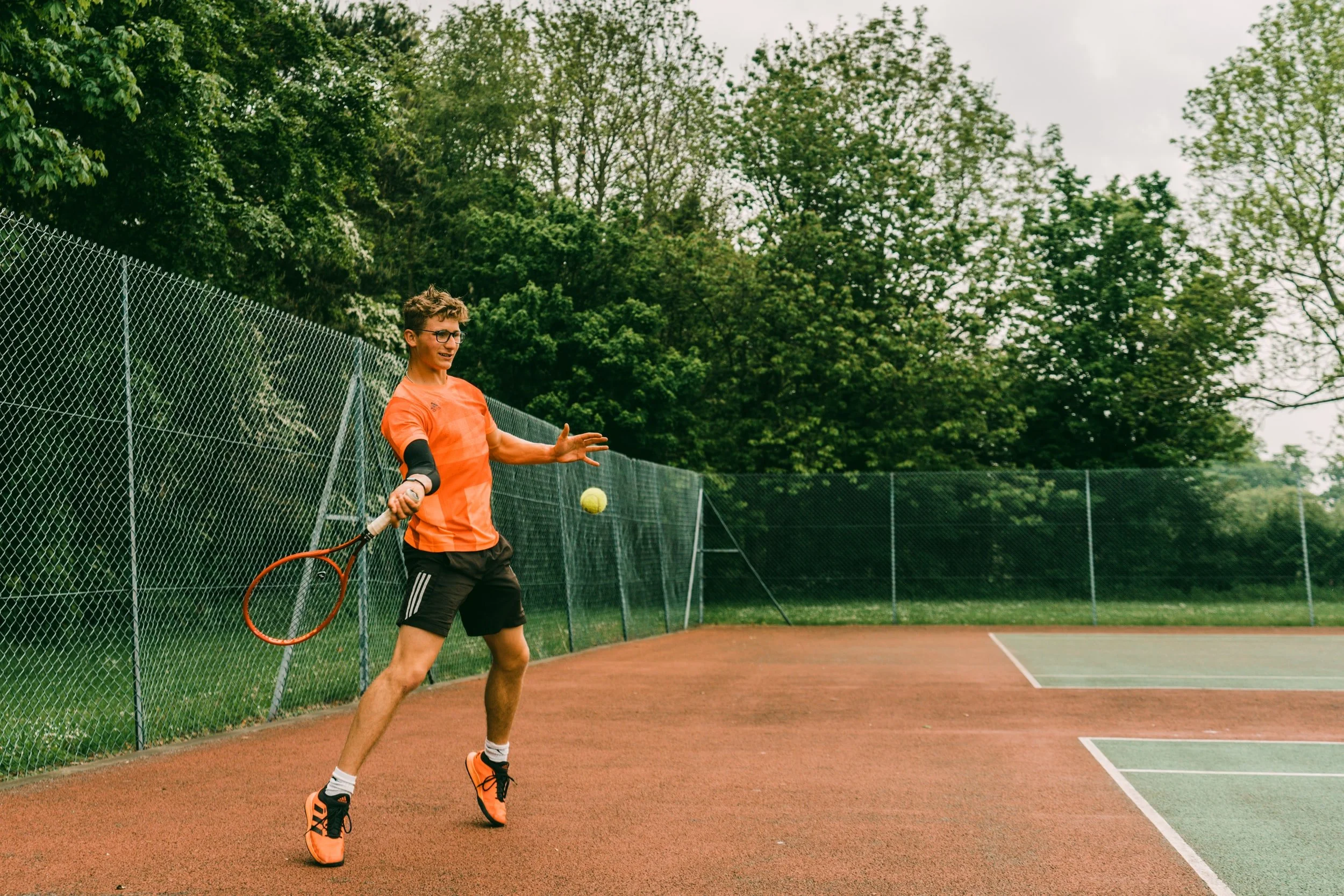 A young man playing tennis on an outdoor court. He is wearing an orange shirt, black shorts, and orange tennis shoes, holding a tennis racket, and appears to be about to hit a tennis ball. There is a green fence around the court and trees in the back
