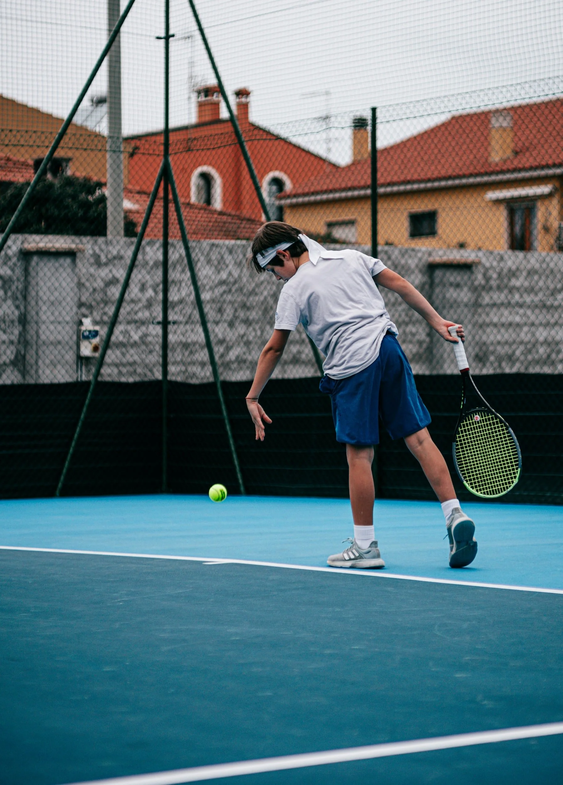 A boy in a white shirt and blue shorts playing tennis on an outdoor court, preparing to hit a tennis ball.