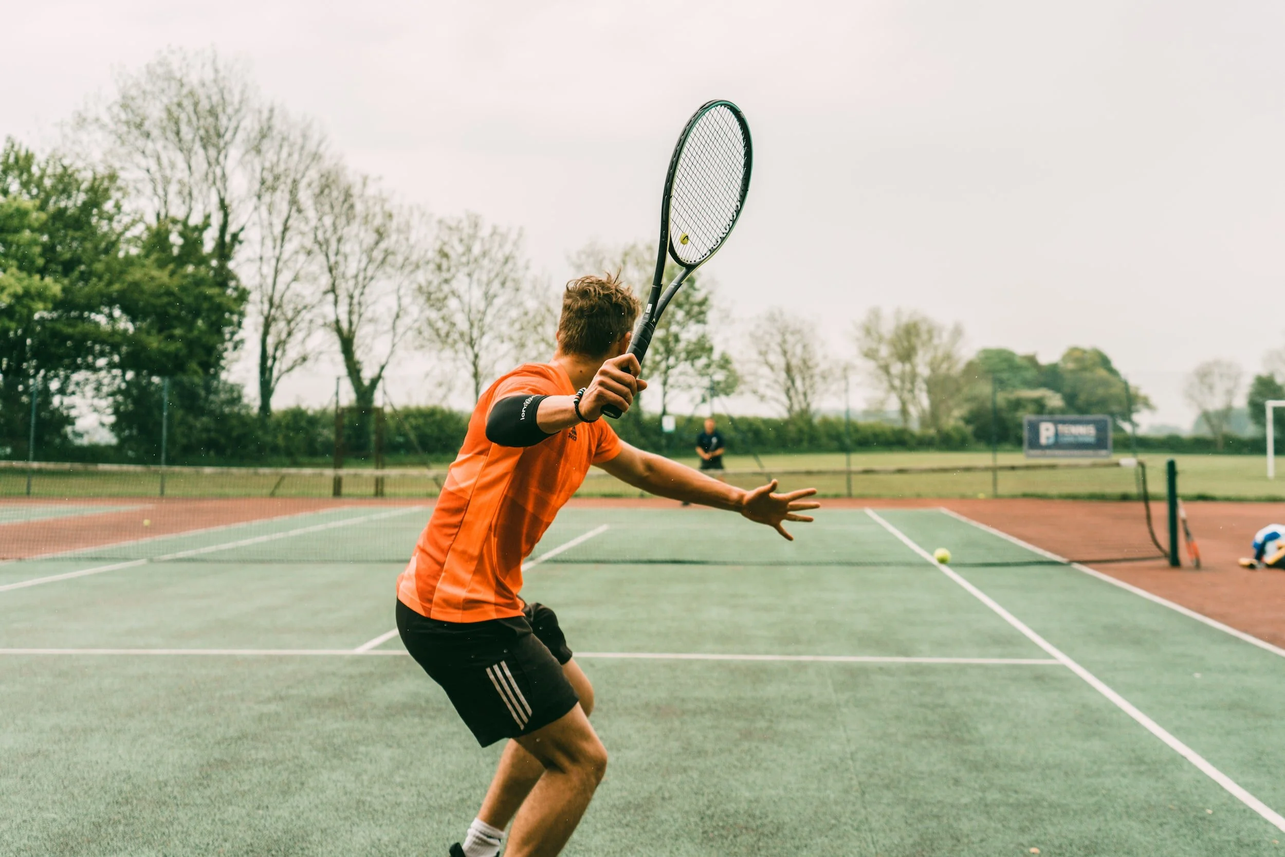 A person playing tennis on an outdoor court, preparing to hit a tennis ball with a racquet, while another person is visible in the background.
