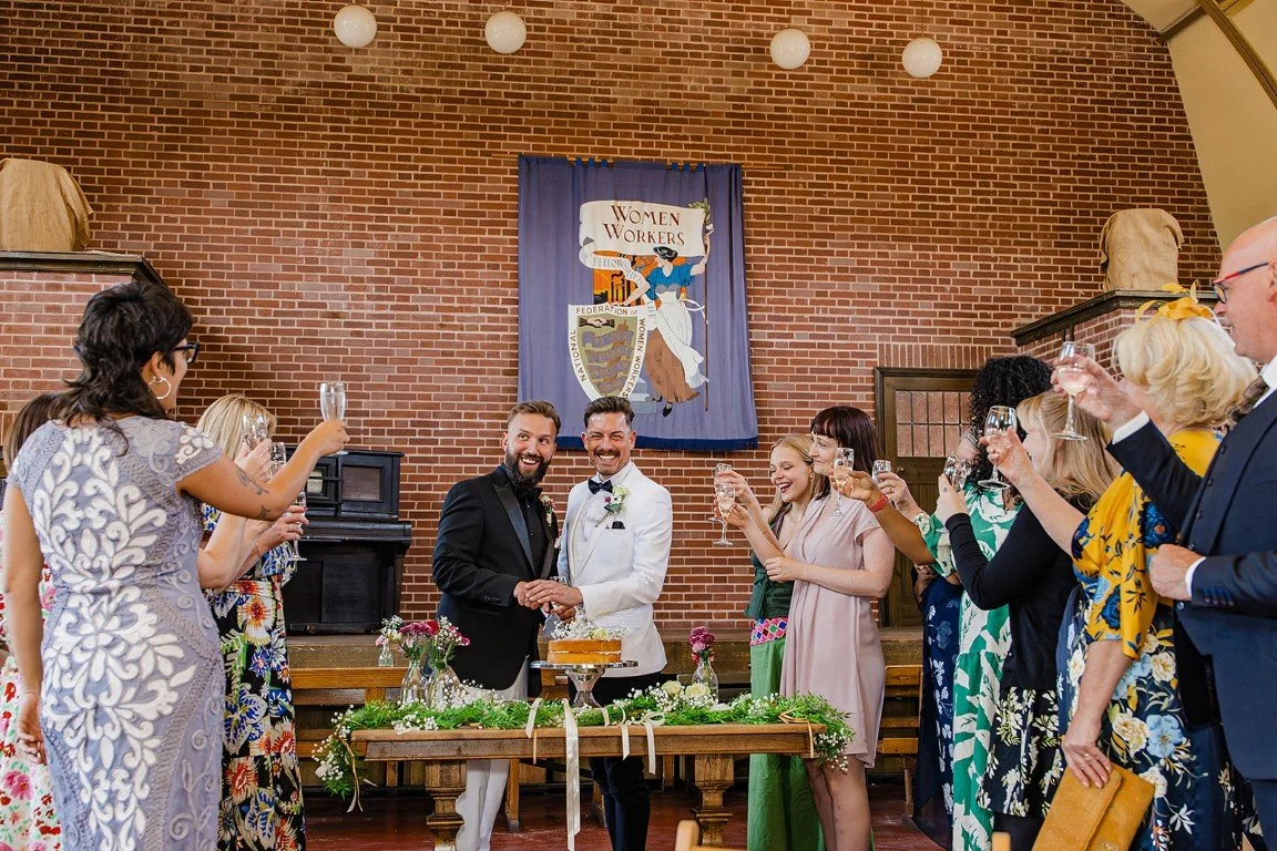 Two men in tuxedos are getting married and are holding hands, smiling at each other, during a wedding celebration. They are surrounded by guests raising champagne glasses in toast, in a room with a brick wall background and a Women's Workers banner h