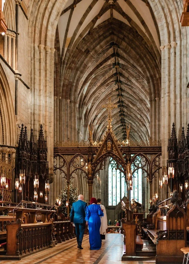 Inside a Gothic-style cathedral with high vaulted ceilings, ornate woodwork, and stained glass windows. Two people, dressed in formal attire, walk down the aisle.