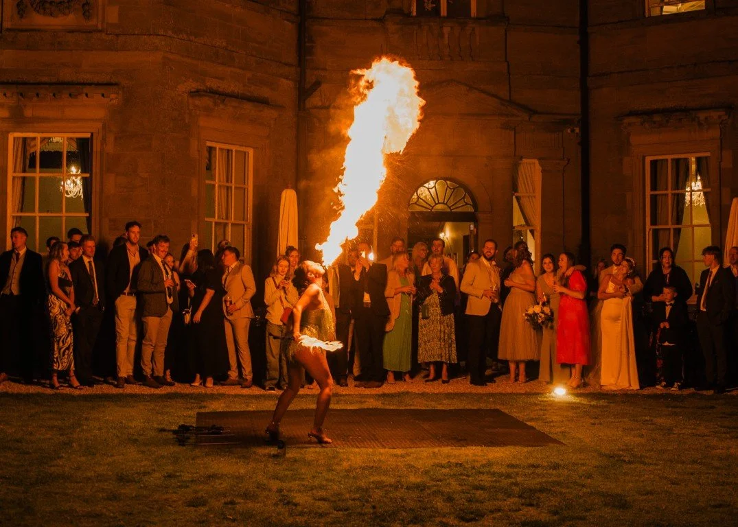 Fire performer at a wedding or party, surrounded by guests, blowing fire into the night sky.