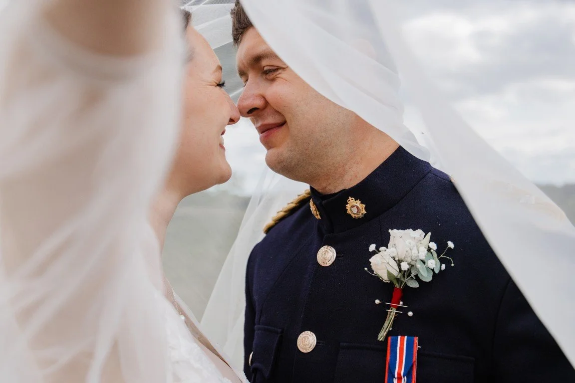 A bride and groom with noses touching under a wedding veil, outdoors on a cloudy day.