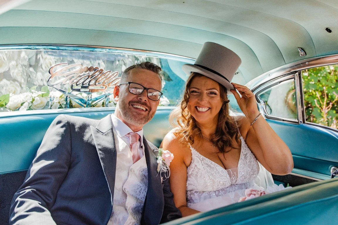 A newlywed couple sitting in the back of a vintage car, smiling and looking at the camera. The bride is wearing a white lace wedding dress and a gray top hat, while the groom is in a gray suit with a pink tie. The car's interior is light blue, and th