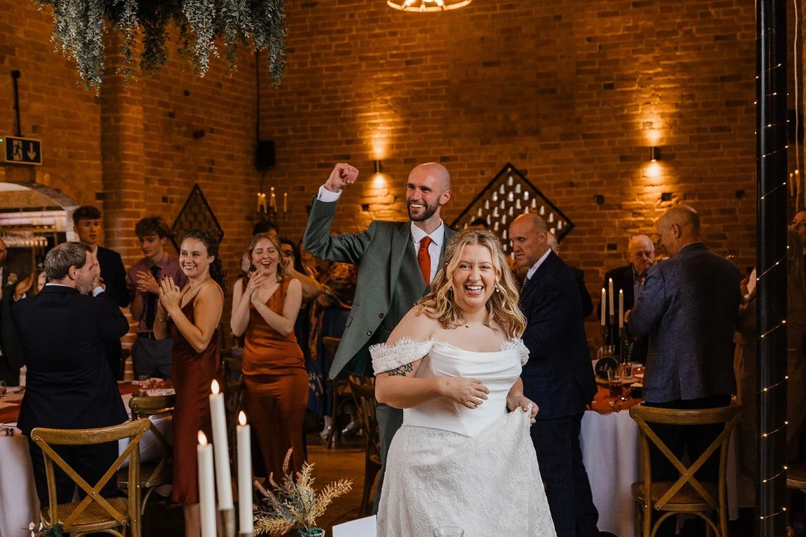 A wedding celebration with the bride and groom dancing and smiling, surrounded by guests clapping and enjoying the moment in a warmly lit venue with brick walls and candlelit tables.