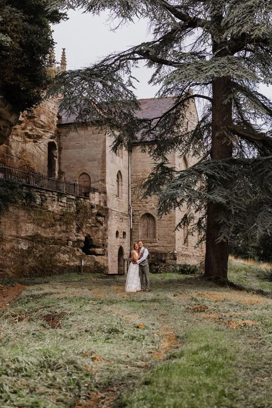 A bride and groom embracing under a tree with Guys Cliff house and chapel in the background