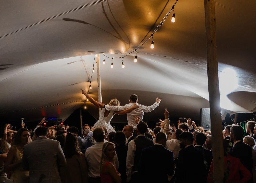 Wedding reception with bride and groom dancing on a chair surrounded by guests, under a canopy decorated with string lights.