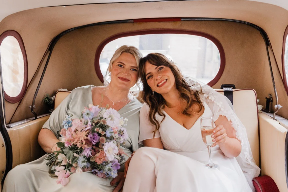 Two women sitting in the back of a vintage van, one in a wedding dress holding a glass of champagne, the other in a pastel-colored dress holding a bouquet of flowers, smiling at the camera.