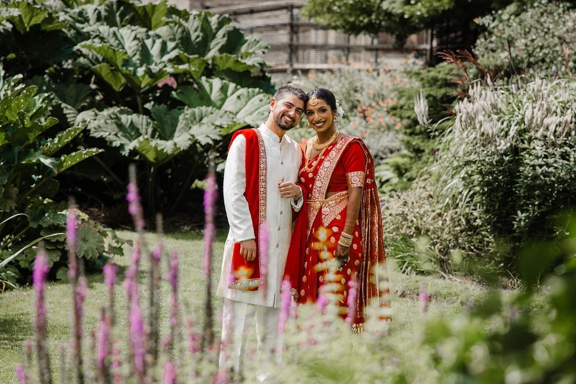A happy couple dressed in traditional Indian wedding attire standing in a lush garden with flowering plants.
