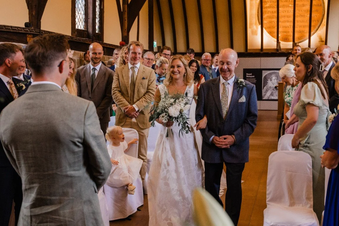 A bride walking down the aisle with her father at her wedding inside a church with guests standing on both sides.