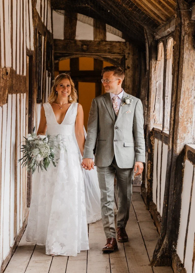 A bride and groom holding hands and walking through a rustic indoor hallway, the bride in a white wedding dress holding a bouquet, and the groom in a light gray suit with a boutonniere.