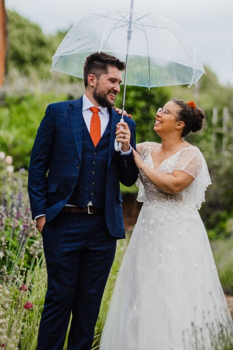 A couple in wedding attire sharing a moment under a clear umbrella outdoors, with greenery in the background.