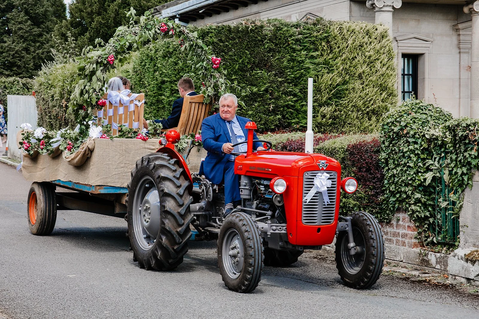Man driving a red tractor pulling a decorated cart with seated people in formal attire, during a wedding procession