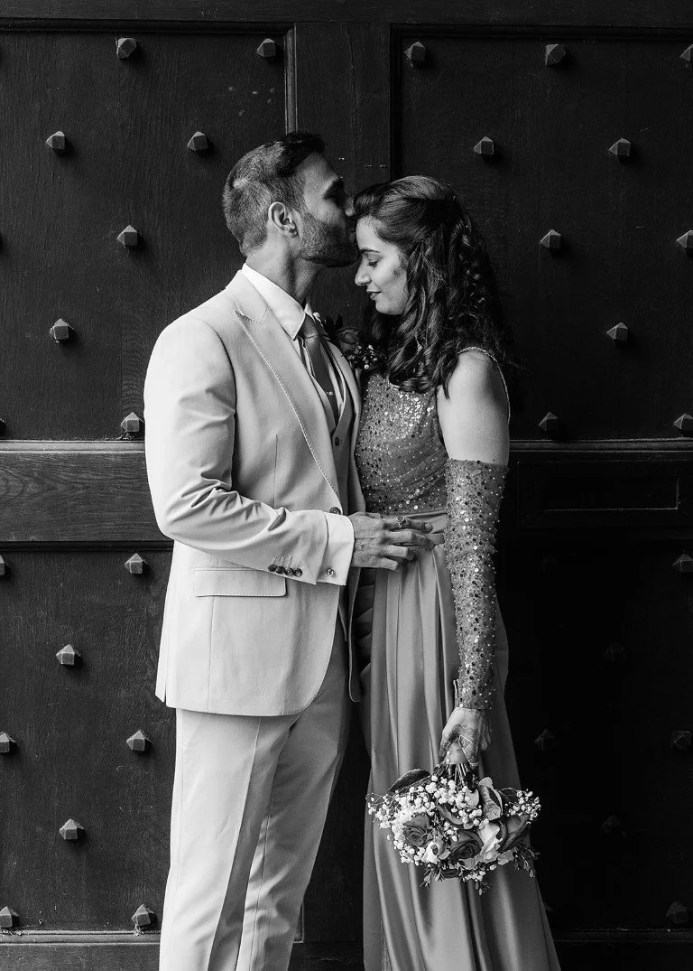 A black and white photo of a couple, the man in a light-colored suit, kissing the woman's forehead. The woman wears a sparkly dress and holds a bouquet of flowers, standing against a textured dark background.