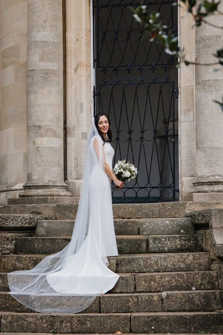 A bride in a white wedding dress with a long veil standing on outdoor stone steps in front of a large metal gate, holding a bouquet of white flowers and smiling.