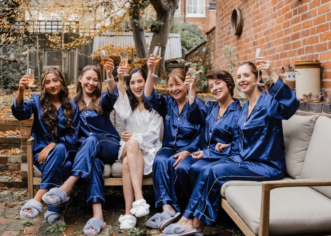Group of six women sitting on an outdoor sofa, celebrating with champagne glasses, wearing matching blue pajamas and slippers, in a backyard with autumn leaves and trees.