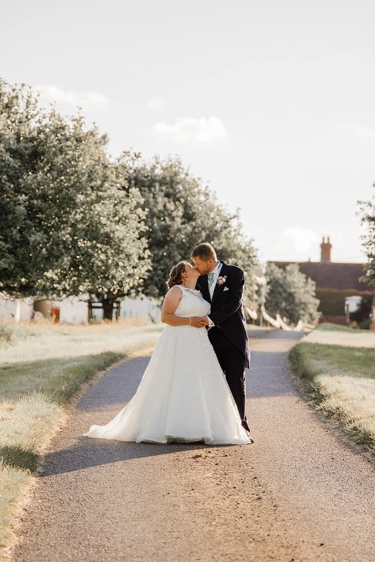 A bride and groom sharing a kiss on a rural road during their wedding, surrounded by trees and a clear sky.